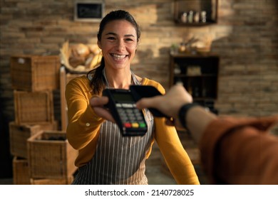 Cinematic shot of young friendly saleswoman passing pos terminal over counter to customer paying with smartphone using NFC technology in bakery shop. - Shutterstock ID 2140728025