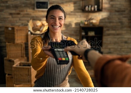 Cinematic shot of young friendly saleswoman passing pos terminal over counter to customer paying with smartphone using NFC technology in bakery shop.