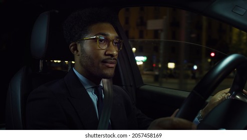 Cinematic Shot Of Young Handsome Afro American Successful Elegantly Dressed Businessman With Glasses Is Driving Modern Luxury Car In Center Of The City By Night.