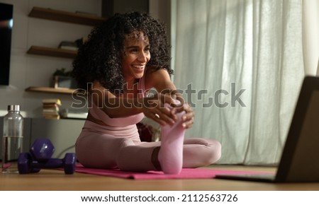 Cinematic shot of young sporty brazilian woman with athletic body wearing bra and leggings is practicing workout exercises for legs and buttocks on fitness mat with online tutorial on laptop at home.