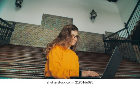 Cinematic Shot Of Young Woman In Yellow Sweater Sit On Stairs Of Old University Campus Building, Work On Laptop On New Exciting School Project Or Assignment. Freshmen Or Junior Student