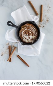 Cinnamon Bun In A Black Skillet Surrounded By Cinnamon Sticks.