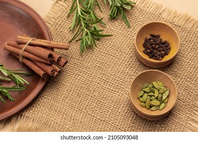 Cinnamon sticks on ceramic plate. Cardamom and cloves in wooden bowls. Sprigs of rosemary. Burlap linen on table. Top view.  - Powered by Shutterstock