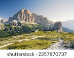 Cinque Torri peaks and Tofane Mountains at summer cloudless morning with green grass lighten by warm light of sun from right side and path inf front