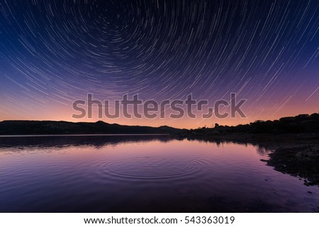 Circle of ripples on a mirror calm lac de Codole in the Balagne region of Corsica at sunrise with dramatic star trails in the pink and purple dawn sky