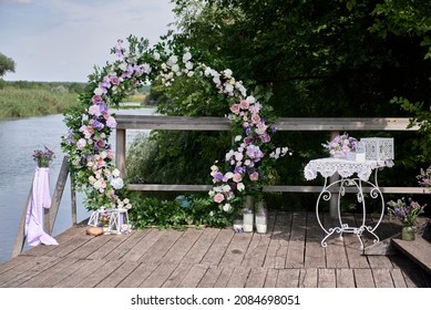 Circle wedding arch decorated with white, pink and purple flowers and greenery on terrace by lake outdoors, copy space. Wedding setting. Floral composition