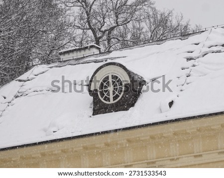 Circular attic window on a snow covered sloped roof of a classical building.
