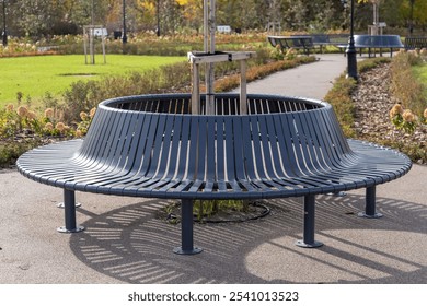 A circular black metal bench encircling a young tree, placed in a green park area, providing ample seating space with a modern design - Powered by Shutterstock