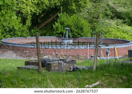 A circular brick structure, possibly a well or cistern, sits partially sunken in a grassy area shaded by trees. Trefor - Gwynedd - Lleyn Peninsula - Wales - Great Britain
