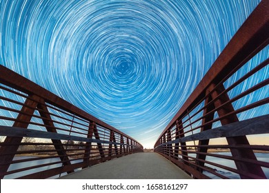 Circular concentric star trails appearing to revolve around Polaris looking North in the night sky, with a wooden boardwalk leading down a bridge. 