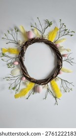 Circular Easter wreath made of intertwined brown vines. Decorated with small branches, vibrant yellow feathers, and pastel pink eggs, including a striped one at the bottom. Delicate green leaves accent the arrangement, set against a light background, creating a festive and natural look.