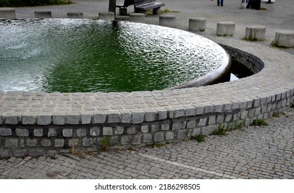 Circular Fountain With A Level Like A Mirror Up To The Top Of The Edge Of The Wall. Water Flowing Over Tiled Cobblestone Granite Wall. Seat Made Of Concrete Cylinder Bench. Circle Around The Fountain.