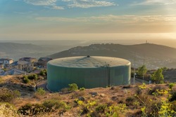 Circular Large Community Water Tank At San Marcos In San Siego, California. Water Tank Near The Slope Of A Hill With Bushes And Shrubs Against The Houses And Mountains At The Back.