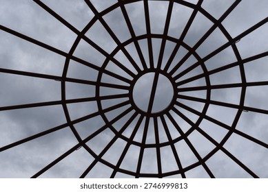 A circular metal framework forms a geometric canopy overhead, with evenly spaced bars radiating outward like spokes from the center. The symmetrical design creates a striking pattern against the cloudy sky, blending structure with open space. This architectural element adds visual interest and modern style while allowing light and air to pass through.