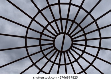 A circular metal framework forms a geometric canopy overhead, with evenly spaced bars radiating outward like spokes from the center. The symmetrical design creates a striking pattern against the cloudy sky, blending structure with open space. This architectural element adds visual interest and modern style while allowing light and air to pass through.