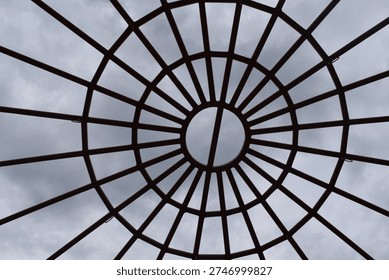 A circular metal framework forms a geometric canopy overhead, with evenly spaced bars radiating outward like spokes from the center. The symmetrical design creates a striking pattern against the cloudy sky, blending structure with open space. This architectural element adds visual interest and modern style while allowing light and air to pass through.