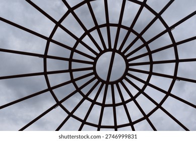A circular metal framework forms a geometric canopy overhead, with evenly spaced bars radiating outward like spokes from the center. The symmetrical design creates a striking pattern against the cloudy sky, blending structure with open space. This architectural element adds visual interest and modern style while allowing light and air to pass through.