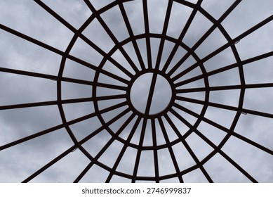 A circular metal framework forms a geometric canopy overhead, with evenly spaced bars radiating outward like spokes from the center. The symmetrical design creates a striking pattern against the cloudy sky, blending structure with open space. This architectural element adds visual interest and modern style while allowing light and air to pass through.