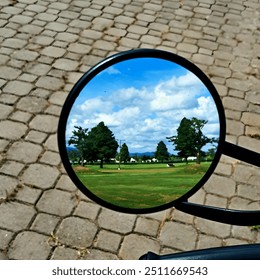 A circular mirror reflects a scenic view of a golf course with green fields, trees, and a blue sky - Powered by Shutterstock