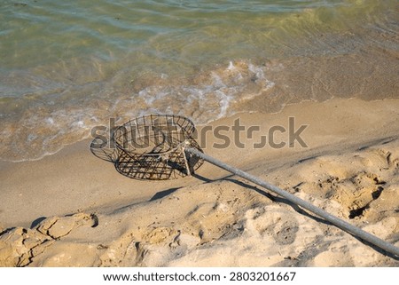 A circular rusty wire mesh basket attached to a weathered wooden pole lies on the wet sand at the edge of the sea, with foamy waves washing up beside it. The abandoned fishing tool casts a geometric shadow on the sand as water approaches, with green algae visible inside the basket and sunlight reflecting off the wet shore around it