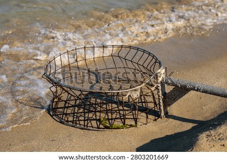 A circular rusty wire mesh basket attached to a weathered wooden pole lies on the wet sand at the edge of the sea, with foamy waves washing up beside it. The abandoned fishing tool casts a geometric shadow on the sand as water approaches, with green algae visible inside the basket and sunlight reflecting off the wet shore around it