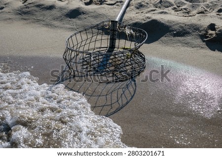 A circular rusty wire mesh basket attached to a weathered wooden pole lies on the wet sand at the edge of the sea, with foamy waves washing up beside it. The abandoned fishing tool casts a geometric shadow on the sand as water approaches, with green algae visible inside the basket and sunlight reflecting off the wet shore around it