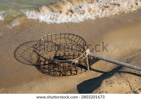 A circular rusty wire mesh basket attached to a weathered wooden pole lies on the wet sand at the edge of the sea, with foamy waves washing up beside it. The abandoned fishing tool casts a geometric shadow on the sand as water approaches, with green algae visible inside the basket and sunlight reflecting off the wet shore around it