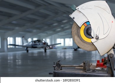 Circular Saw In Front Of Small Single Prop Engine Aircraft Plane Parked Inside Workshop Hangar With Opened Door At Aerodrome. Maintenance Of Private Ultralight