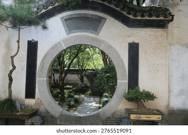 Circular stone arch frames a tranquil garden view. Green trees and a gravel path extend through the arch. Potted bonsai trees sit on either side of the archway