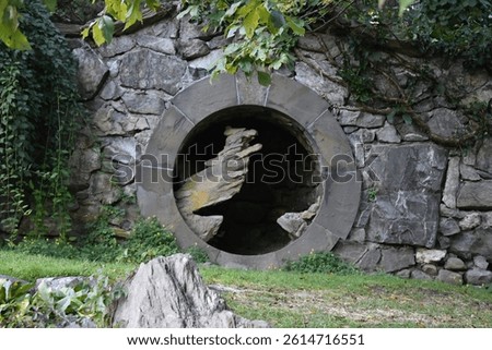 Circular stone portal embedded in a rustic stone wall, with abstract, jagged rock formations emerging from within the tunnel-like opening. Surrounded by creeping ivy and lush greenery.