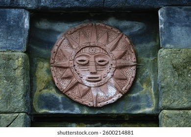 A circular stone relief depicting a sun face with intricate patterns and textures, set against a weathered stone wall. The sun has rays extending outward, showcasing artistic craftsmanship.