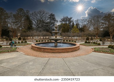 a circular water fountain in the garden surrounded by red brick, bare winter trees and lush green trees with blue sky and powerful clouds at sunset at Memphis Botanic Garden in Memphis Tennessee USA