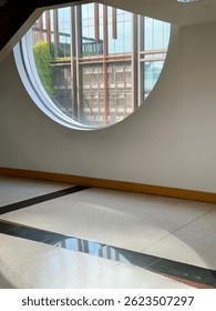 Circular window in a modern interior frames an exterior view of architectural elements and natural light casting shadows on the floor.