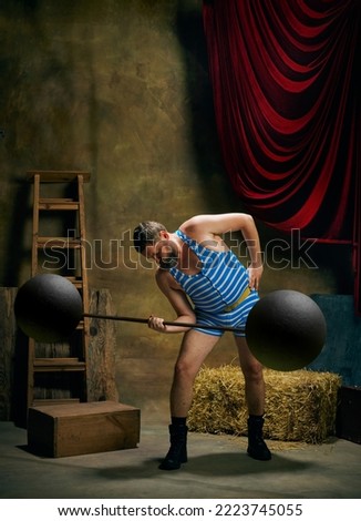 Circus athlete. Vintage portrait of retro circus strongman wearing blue striped sports swimsuit training with barbell over dark circus backstage background. Concept of creative art, fashion, style