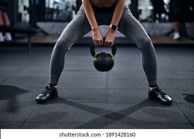 Citizen in grey sport trousers holding the kettlebell between her legs by the handle with both hands