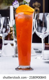 Citrus Fruit, Grapefruit And Lemon Lemonade In A Tall Glass With Ice And An Orange Slice On Top. The Glass Is On A White Tablecloth, In The Back There Are 4 Wine Glasses, Empty Glasses And A Vase Of F