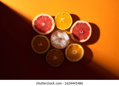 Citrus Fruits Cut In Half And Ice Cubes In Glass On Orange Background, In Sunlight. Preparing Cold Summer Cocktail Concept. Making Tropical Drink