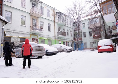 City After Blizzard. Cars Covered With Snow Parked On A Yard, Man Sweeps Snow From His Parked Car, Children Watching. January 12, 2017. Kyiv, Ukraine