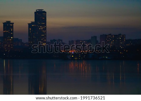 City block on the river bank. Dark silhouettes of buildings with glowing windows are reflected in the water against the setting sun. 