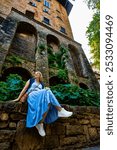  City break in Italy. Beautiful young woman in long blue dress sitting and looking around during sightseeing old city of Bergamo on warm autumn day. Visit old town of Bergamo, Lombardy. Front view.