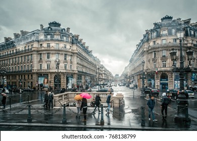 City Center Street/avenue Corner In Paris. People Under Colored Umbrellas Run In The Rain On The Streets Of Paris, France