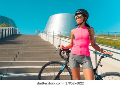 City cyclist woman with road bike. Girl wearing helmet, sunglasses, pink jersey for biking on hot summer day urban commute ride. Cycling concept.