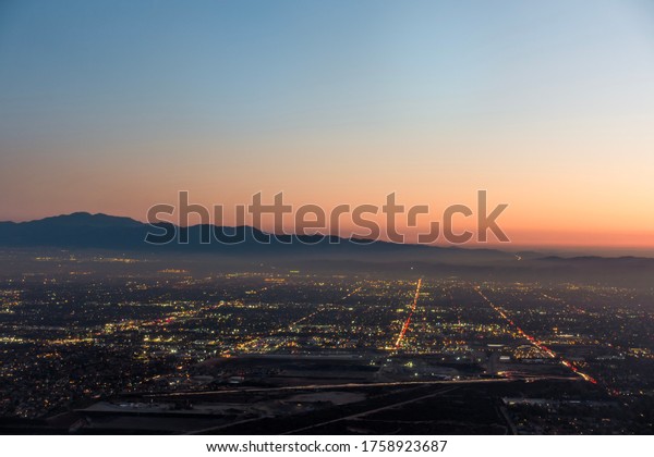 The city lights of the skyline of the Inland Empire\
near Los Angeles California begin to appear as the sun sets in a\
dramatic orange sunset. View from Potato Mountain in Claremont\
Wilderness Park