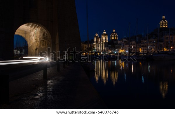 A\
city at night. A night view in Birgu, Malta - waterfront with a\
church and a light trails of a car passing a tunnel\
