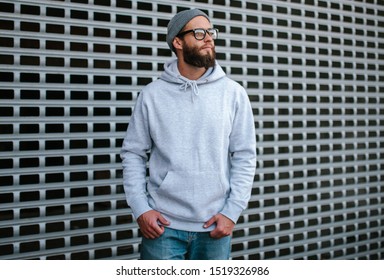 City portrait of handsome hipster guy with beard wearing gray blank hoodie or sweatshirt and hat with space for your logo or design. Mockup for print