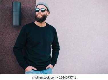 City Portrait Of Handsome Hipster Guy With Beard Wearing Black Blank Hoody Or Sweatshirt And Hat With Space For Your Logo Or Design. Mockup For Print