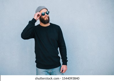 City Portrait Of Handsome Hipster Guy With Beard Wearing Black Blank Hoody Or Sweatshirt And Hat With Space For Your Logo Or Design. Mockup For Print