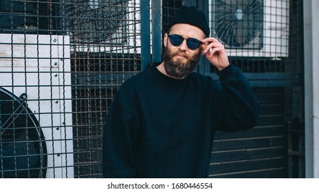 City Portrait Of Handsome Hipster Guy With Beard Wearing Black Blank Hoody Or Sweatshirt  With Space For Your Logo Or Design And Casual Beanie. Mockup For Print