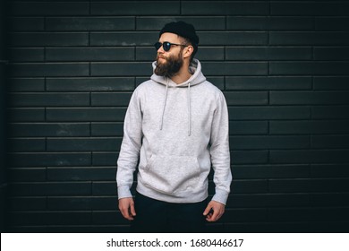 City Portrait Of Handsome Hipster Guy With Beard Wearing Gray Blank Hoodie Or Hoody With Space For Your Logo Or Design And Casual Beanie. Mockup For Print