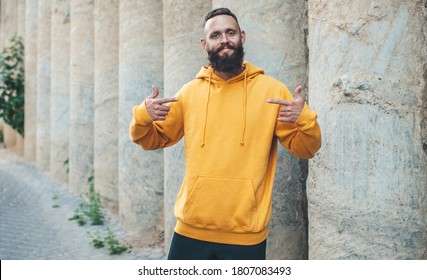 City Portrait Of Handsome Hipster Guy With Beard Wearing Yellow Blank Hoodie Or Sweatshirt With Space For Your Logo Or Design. Mockup For Print